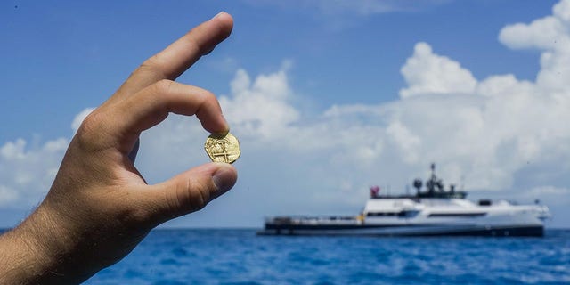 Un explorador sostiene una moneda de oro encontrada en las Bahamas, donde se puede ver desde lejos el barco de exploración de Allen.