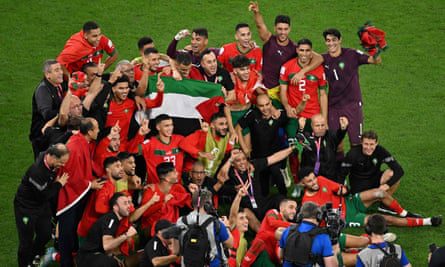 Un grupo de jugadores con sus camisetas rojas se reúnen para una foto en el campo, gesticulando en celebración.  Varios jugadores izan la bandera de Palestina en medio del grupo